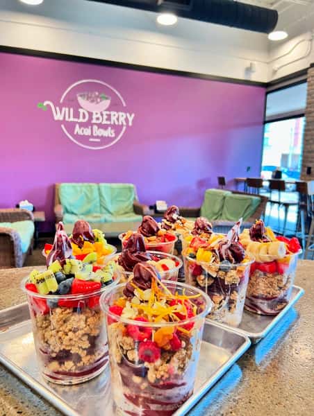 Colorful acai bowls from Wild Berry Acai Bowls, each layered with granola, fresh berries, banana slices, kiwi, and rich acai soft serve, served in clear plastic cups on metal trays inside a modern smoothie café with a vibrant purple accent wall.