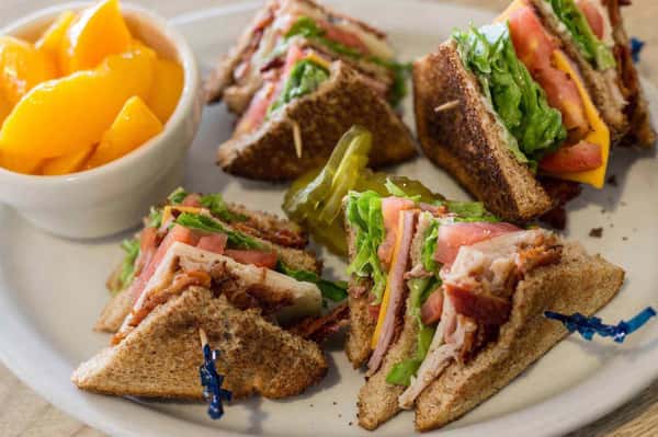 Club sandwich triangles with toasted whole-grain bread, layered turkey, crispy bacon, lettuce, tomato, and cheese, secured with toothpicks and served with a side of peach slices—classic American diner lunch plate.