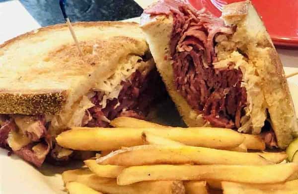 Hot pastrami sandwich on toasted bread with melted cheese and sauerkraut, served with crispy French fries—classic deli-style Reuben sandwich plate.