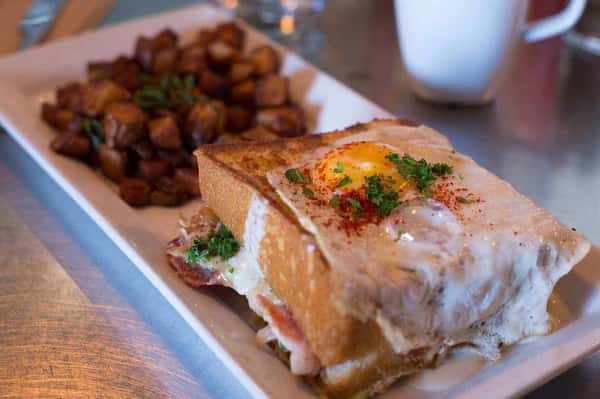 Croque madame sandwich topped with creamy béchamel and a fried egg, served with crispy breakfast potatoes—French-inspired brunch dish.