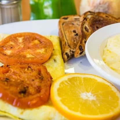 Classic diner breakfast with omelet, grilled tomato slices, raisin toast, orange slice, and creamy grits.