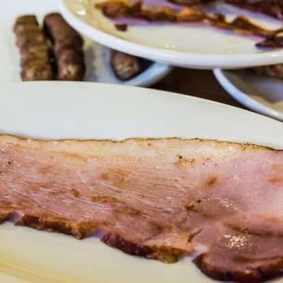 Close-up of sliced roasted ham on a white plate with breakfast sausages and bacon in the background at a classic diner.