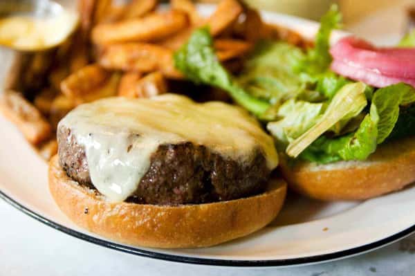 Juicy cheeseburger with melted white cheese on a toasted bun, served open-faced with fresh lettuce and pickled onions, alongside crispy seasoned fries and dipping sauce—classic American restaurant burger plate.