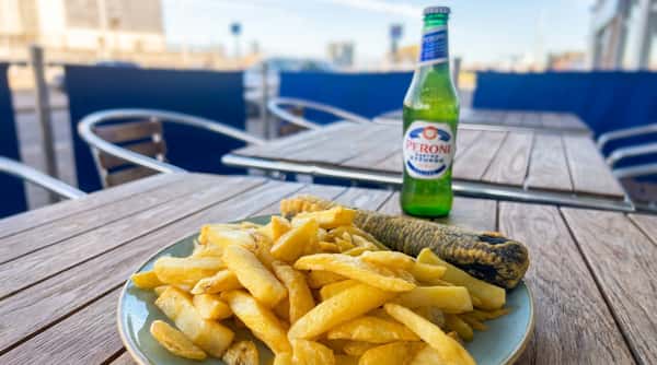 Plate of golden french fries and a dark battered sausage on a wooden table with a Peroni beer bottle.