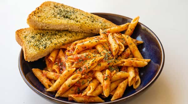 Penne pasta with red sauce and green herbs, served in a dark bowl with two slices of garlic bread.