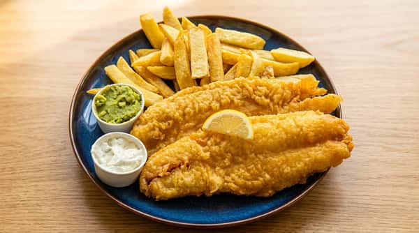 Fish and chips with two battered fish fillets, golden french fries, a lemon wedge, tartar sauce, and mushy peas on a blue plate.