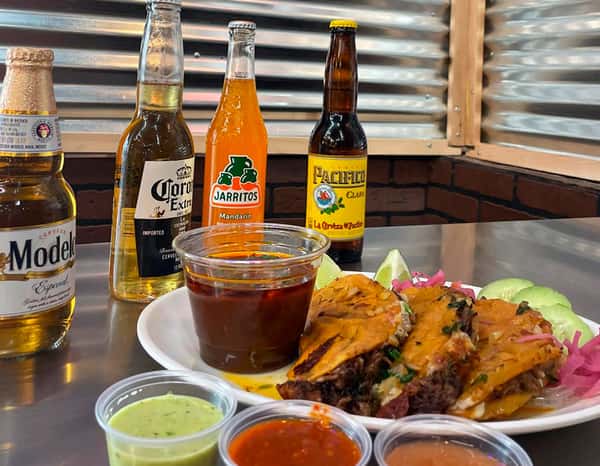Authentic Mexican birria tacos served with rich consommé, fresh cucumber slices, and pickled onions, paired with Jarritos mandarin soda and Pacifico beer at Tacos Chano, accompanied by three flavorful dipping salsas.