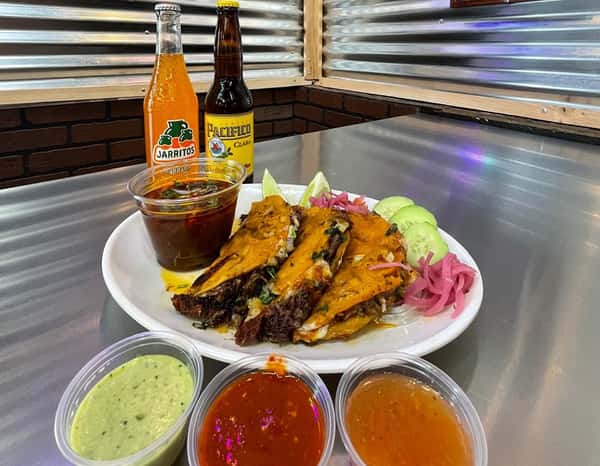 Authentic Mexican birria tacos served with rich consommé, fresh cucumber slices, and pickled onions, paired with Jarritos mandarin soda and Pacifico beer at Tacos Chano, accompanied by three flavorful dipping salsas.
