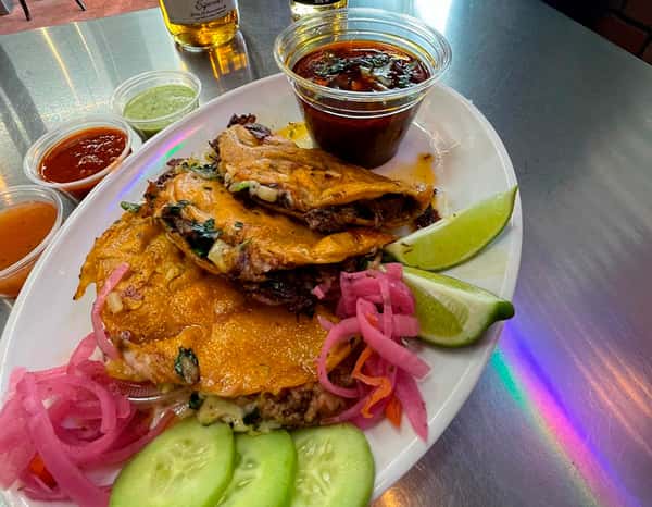 Authentic birria tacos filled with slow-cooked beef, melted cheese, and cilantro, served with rich consommé, lime wedges, pickled onions, and fresh cucumber slices at Tacos Chano.