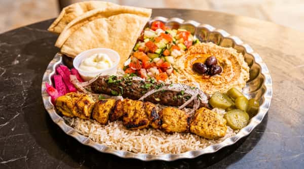 Kebab platter with chicken and kofta skewers on rice, hummus, salad, pita bread, pickles, and garlic sauce.