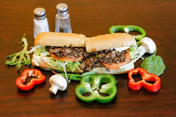 Steak hoagie sandwich with shredded lettuce, tomato, and beef, surrounded by bell peppers, mushrooms, and greens on a wooden table.