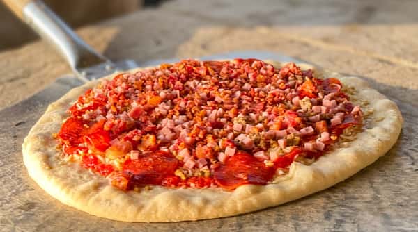 Uncooked pizza with pepperoni, diced ham, and bacon crumbles on a white dough in a pizza oven.