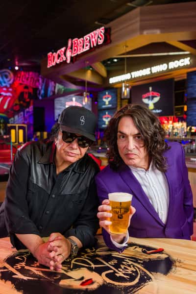 Gene Simmons and Paul Stanley of KISS pose inside Rock & Brews, with Paul Stanley holding a Rock & Brews craft beer at the bar while Gene Simmons leans forward, surrounded by the restaurant's rock-themed décor and illuminated signage.