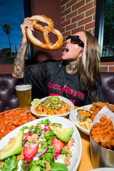Tattooed woman in sunglasses holds up an oversized pretzel at Rock & Brews, surrounded by pizza, nachos, salad, chicken tenders, fries, wings, and beer.