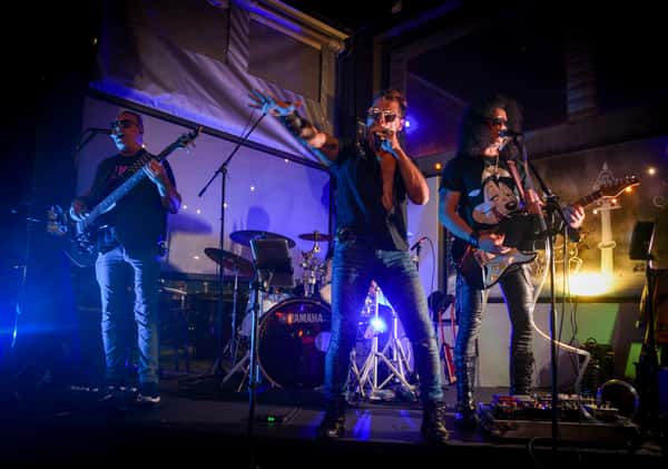 A rock band performs live on an illuminated stage, featuring a lead singer gesturing to the crowd, a bassist, a guitarist, and a drum kit under blue and white stage lighting.