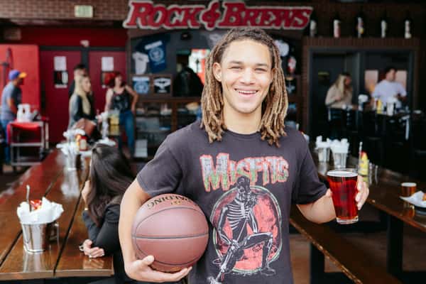 A smiling man stands in a Rock & Brews restaurant, wearing a Misfits band T-shirt. He holds a basketball under one arm and a glass of dark beer in his other hand. Long communal wooden tables and restaurant patrons are visible in the background, creating a casual, upbeat atmosphere.