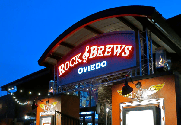 A vibrant exterior shot of the Rock & Brews restaurant in Oviedo at dusk. The entrance features a large, arched sign illuminated in red and white neon against a deep blue sky. The building has bright orange walls, rock-and-roll-themed logos, and warm outdoor lighting with decorative string lights and fire features.
