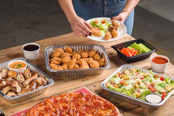 Catering for a large group of guests seated at a long banquet-style table enjoying a meal with private dining event in Oviedo, FL. The table has a spread with Pretzels, Boneless Wings, Salad, and Pizzas