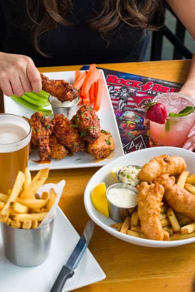 Close-up of scratch-made sweet & spicy asian wings, fish and chips, fries, and a strawberry basil margarita on a Rock & Brews table, highlighting classic American comfort food served in a rock-and-roll–inspired restaurant.