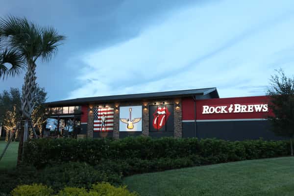 The exterior of the Rock & Brews Orlando restaurant featuring a modern building with rock-themed murals including the American flag, a winged guitar, and the Rolling Stones lips logo. The restaurant's name is displayed in large white letters against a red and black wall, set against a backdrop of a cloudy twilight sky and green landscaping.