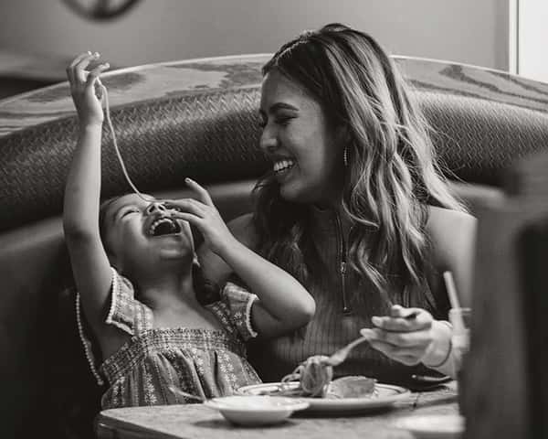 A young girl feeds a long noodle into her mouth while laughing