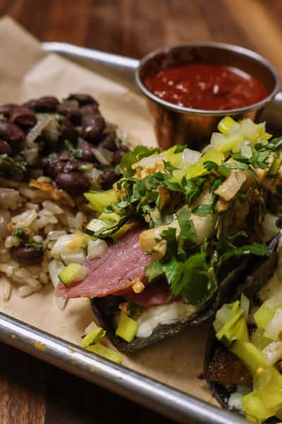 Taco filled with pastrami, melted cheese, onions, and cilantro on a blue corn tortilla, served with a side of salsa.