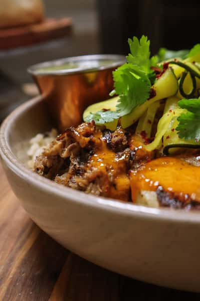Shredded meat with melted cheese, zucchini ribbons, and cilantro served over rice in a bowl with a copper sauce cup.