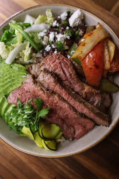 Steak bowl with sliced grilled beef, avocado, black beans, sautéed peppers and onions, lettuce, zucchini, and queso fresco.