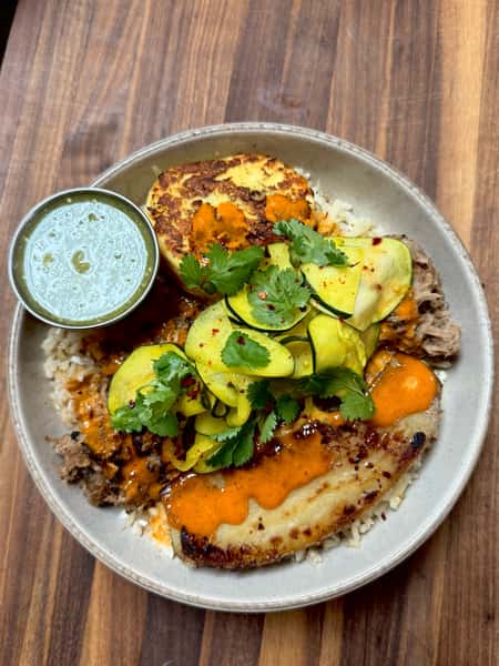 Pork and rice bowl with fried plantain, squash ribbons, cilantro, and spicy orange sauce, served with a green dipping sauce.