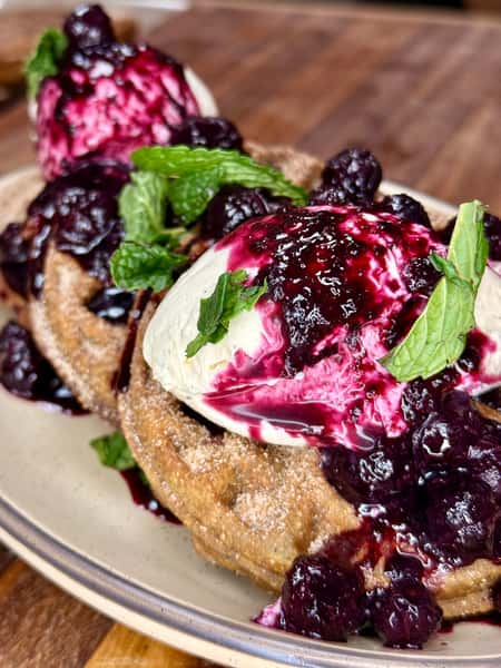 This image showcases a delicious dessert dish featuring crispy waffles dusted with cinnamon sugar, generously topped with vanilla ice cream or whipped cream, a vibrant berry compote (likely blueberry), and fresh mint leaves for garnish. The contrast between warm waffles and cold cream, plus the sweet and tart berries, makes it visually and flavorfully indulgent.