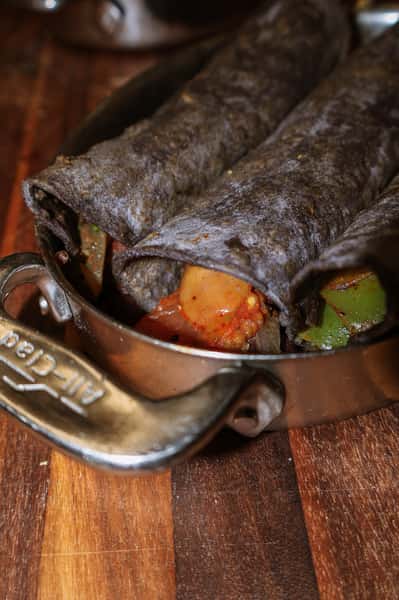 Blue corn tortillas rolled and filled, likely enchiladas or taquitos, with visible roasted vegetables and beans peeking out—served in a metal skillet.