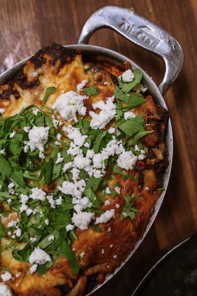 Baked casserole topped with melted cheese, fresh cilantro, and crumbled queso fresco in a metal skillet on a wooden surface.