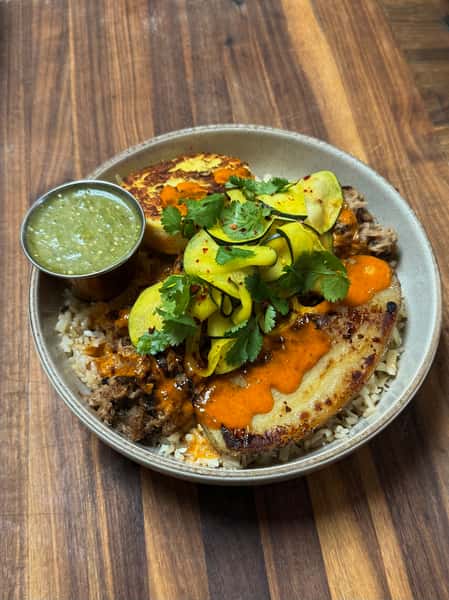 Pork and rice bowl with fried plantain, squash ribbons, cilantro, and spicy orange sauce, served with a green dipping sauce.