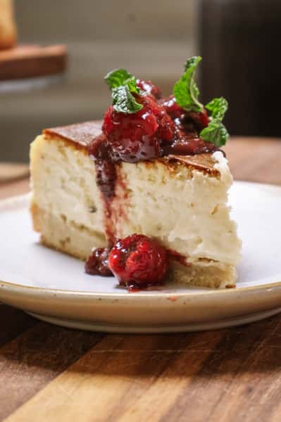 Slice of cheesecake topped with berry compote and fresh mint on a speckled ceramic plate.