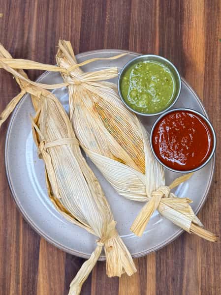 Three corn husk-wrapped tamales served with green tomatillo salsa and red chili sauce on a ceramic plate.
