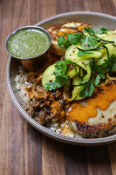 Pork and rice bowl with fried plantain, squash ribbons, cilantro, and spicy orange sauce, served with a green dipping sauce.