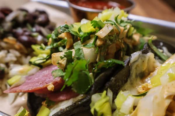 Taco filled with pastrami, melted cheese, onions, and cilantro on a blue corn tortilla, served with a side of salsa.