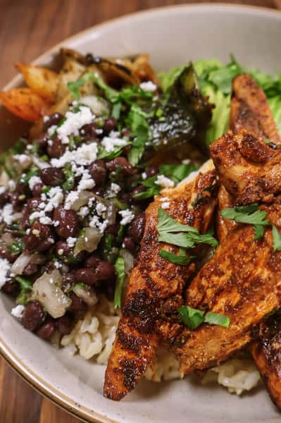 Grilled chicken bowl with seasoned rice, black beans, sautéed vegetables, lettuce, and queso fresco, topped with fresh cilantro.