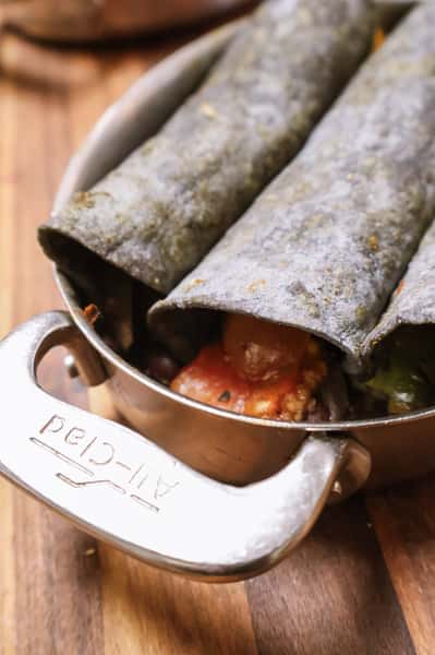 Blue corn tortillas rolled and filled, likely enchiladas or taquitos, with visible roasted vegetables and beans peeking out—served in a metal skillet.