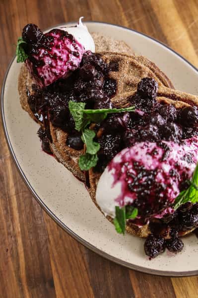 This image showcases a delicious dessert dish featuring crispy waffles dusted with cinnamon sugar, generously topped with vanilla ice cream or whipped cream, a vibrant berry compote (likely blueberry), and fresh mint leaves for garnish.