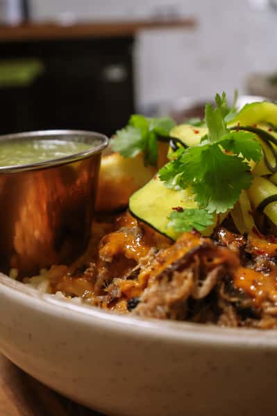 Pork and rice bowl with fried plantain, squash ribbons, cilantro, and spicy orange sauce, served with a green dipping sauce.