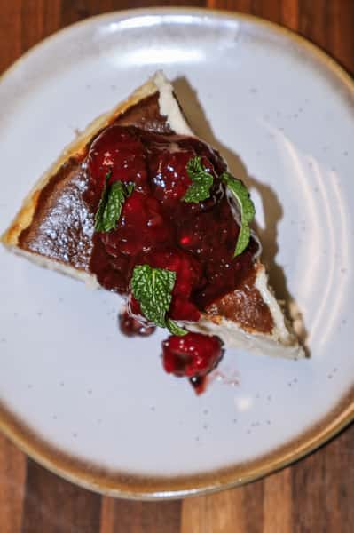 Slice of cheesecake topped with berry compote and fresh mint on a speckled ceramic plate.