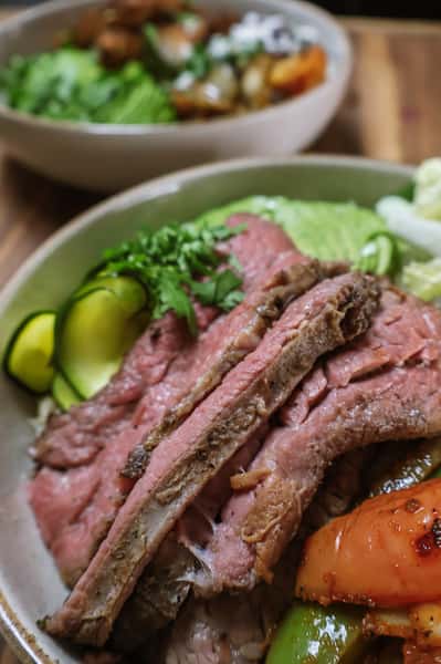 Sliced grilled steak served over rice with avocado, sautéed peppers, black beans, greens, and zucchini ribbons in a bowl.