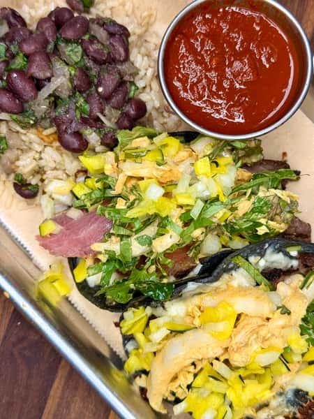 Two blue corn tacos topped with shredded lettuce, onions, and diced pickles, served with rice, black beans, and a side of red salsa.
