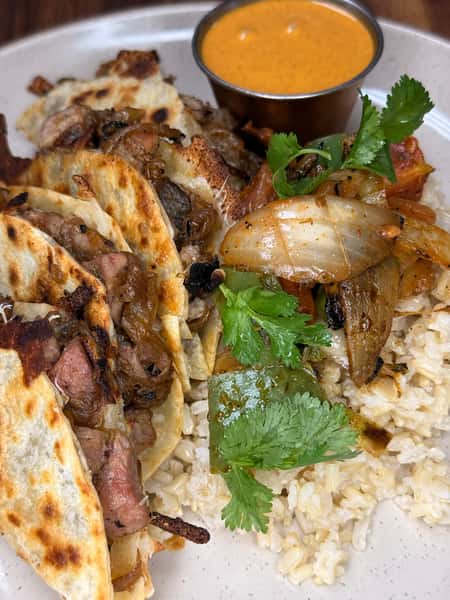 Grilled steak tacos with sautéed onions and cheese, served with rice, roasted vegetables, fresh cilantro, and creamy dipping sauce.