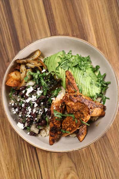 Grilled chicken bowl with sliced avocado, black beans, rice, sautéed peppers and onions, queso fresco, and fresh cilantro on a wooden table.