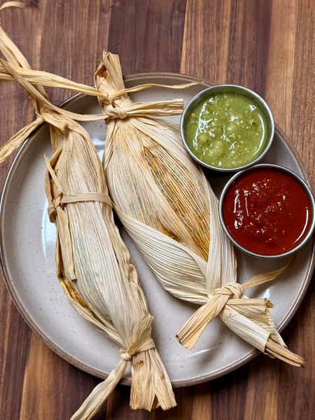 Three corn husk-wrapped tamales served with green tomatillo salsa and red chili sauce on a ceramic plate.