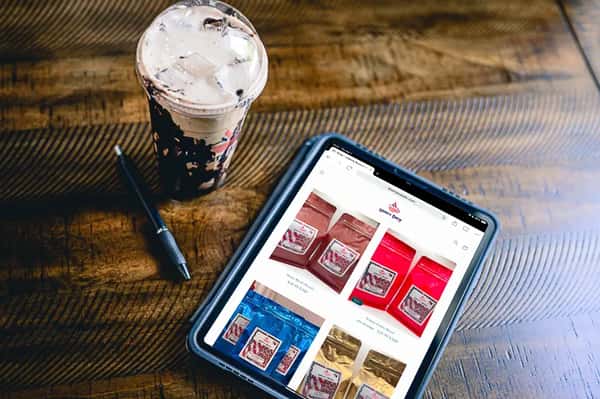 Iced mocha drink with chocolate drizzle beside a tablet displaying Liberty Brew's online coffee shop, featuring various patriotic-themed coffee bag blends, on a rustic wooden table.