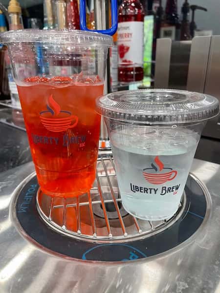 Two Liberty Brew cold drinks—iced cherry refresher and ice water—in branded clear plastic cups, displayed on a modern drink prep station.