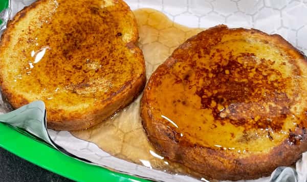 Thick-cut French toast slices drenched in maple syrup, served in a green basket lined with parchment paper for a comforting breakfast option.